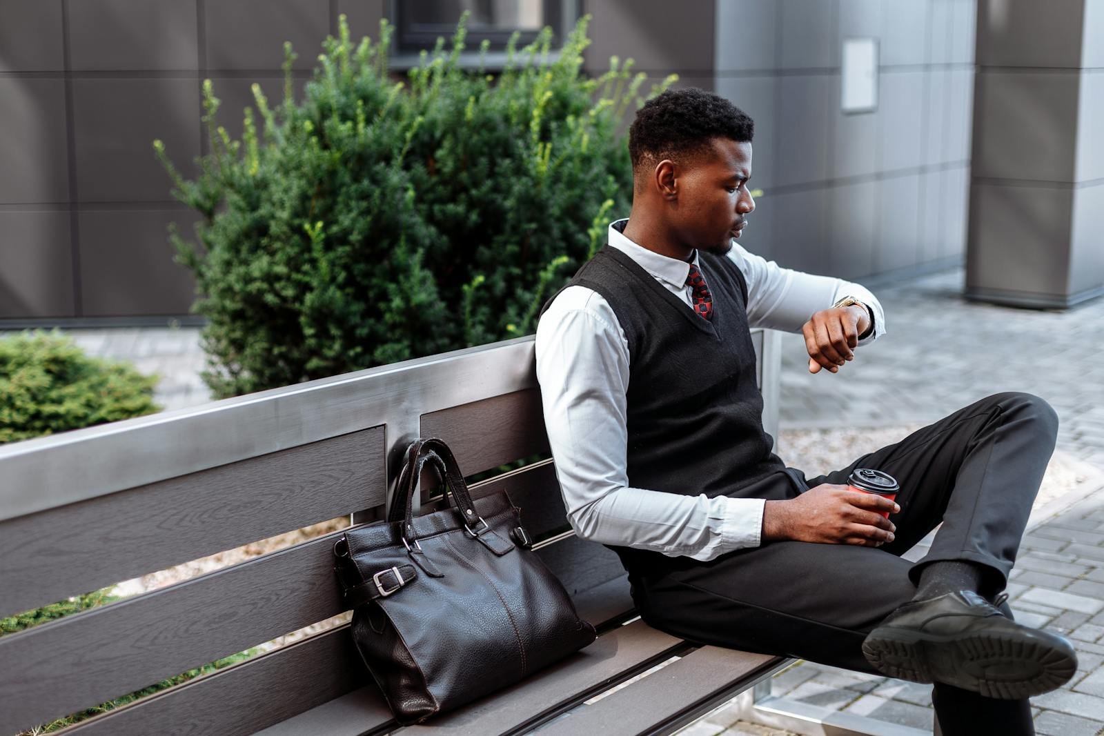 Young professional sitting on a bench, checking his wristwatch with coffee in hand.