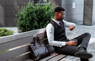 Young professional sitting on a bench, checking his wristwatch with coffee in hand.
